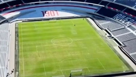 Vista aérea del campo de juego del estadio Monumental, mostrando sectores con césped irregular y arena visible.